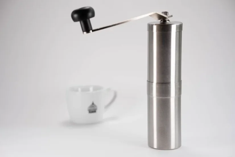Strieborný ručný mlynček na biele pozadí so šálkou kávy