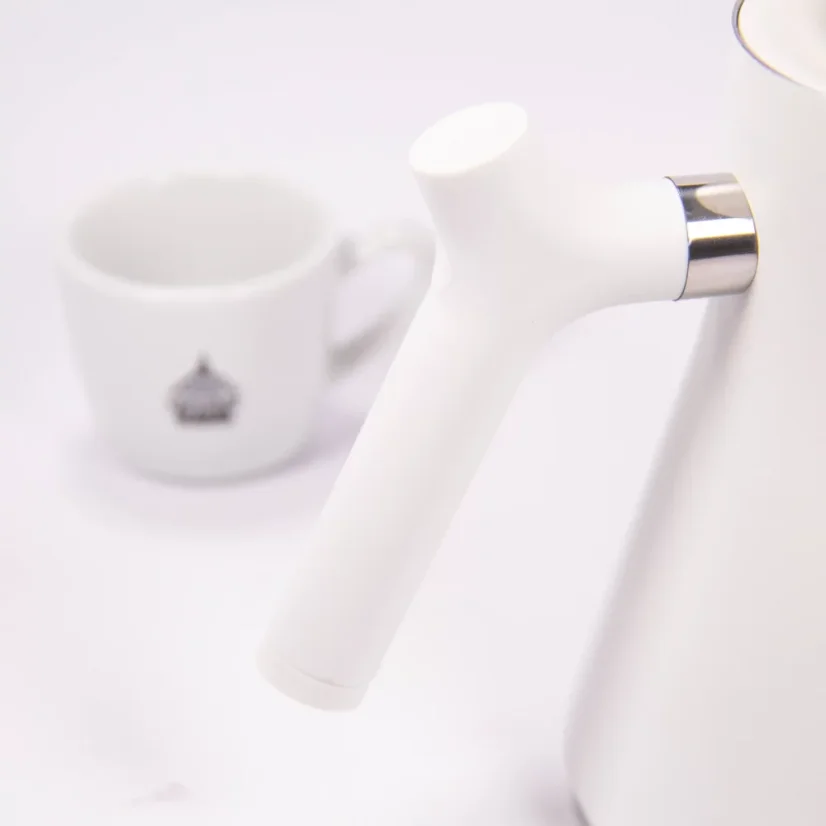 Biela elektrická kanvica, detail na rukoväť kanvice, v pozadí šálka kávy.
