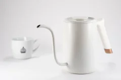Weiße Schnellkochkanne der Marke Timemore Fish Pure Over auf weißem Hintergrund zusammen mit einer Tasse mit Motiven. Weiße Schnellkochkanne der Marke Timemore Fish Pure Over auf weißem Hintergrund zusammen mit einer Tasse mit Motiven.