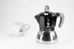 Cafetera moka de aluminio adecuada para inducción con logo del fabricante - la empresa italiana Bialetti, en composición con una taza con logo.