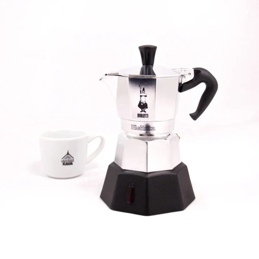 Strieborná moka kanvica so zdrojom ohrevu značky Bialetti Moka Elettrika Standard s šálkom kávy v pozadí.