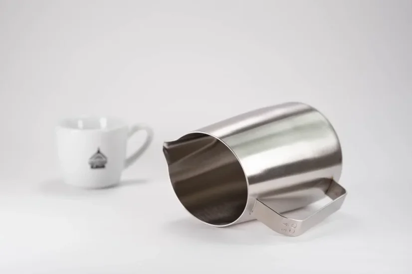 Symmetrische Kanne aus Edelstahl zur Zubereitung von aufgeschäumter Milch mit einem Volumen von 600 ml auf einem Tisch mit einer weißen Tasse Kaffee.