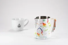 White stainless steel milk frothing jug with colorful splashes from Barista Space Splash, accompanied by a cup bearing a coffee logo.