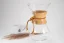 Chemex en verre avec poignée en bois et lanière en cuir, tasse à café blanche et café moulu dispersé sur une table blanche.