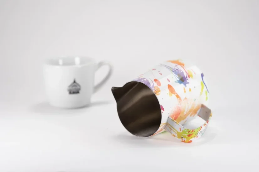 Detail of the inner wall of a stainless steel milk frothing pitcher in white with colorful design accents from the Barista Space Splash brand, featuring a cup with a logo.
