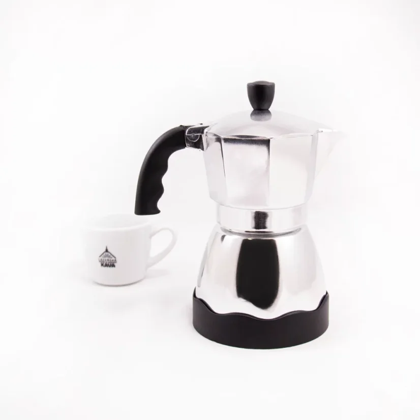 Side view of a silver Bialetti Moka Timer coffee maker with a white porcelain cup with motifs in the background.
