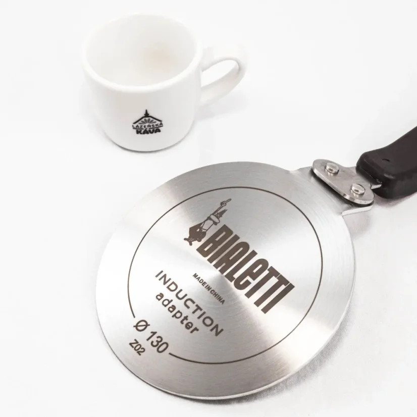 Detail of the back side of a Bialetti induction plate on a table, accompanied by a white cup with coffee motifs.