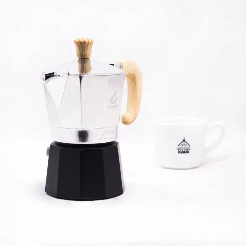 Strieborná moka konvička s drevenou rukoväťou značky Forever Miss Moka Woody pre 3 porcie kávy s porcelánovým šálkom na pozadí.