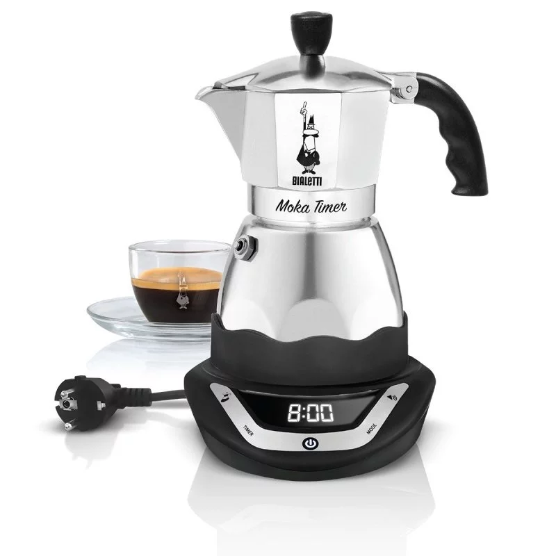 Silver Bialetti Moka Timer coffee maker for 3 cups, placed on a black stand with a glass cup of prepared coffee in the background.