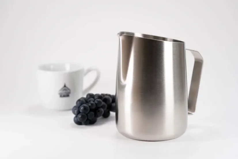 Symmetrical stainless steel milk frothing pitcher placed on a table alongside red grapes and a cup with coffee-themed patterns.