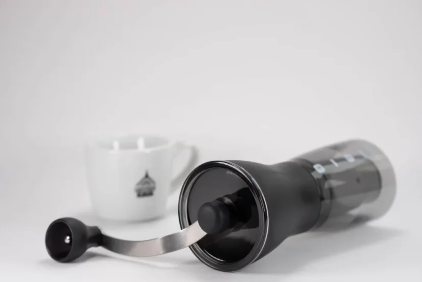 Čierny ručný mlynček na kávu ležiaci na biele stole a biely šálka na kávu v pozadí.