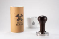 Heavy Temper with a diameter of 58 mm and a black handle on a white table with a cup of coffee and the original packaging for the temper. Heavy Temper with a diameter of 58 mm and a black handle on a white table with a cup of coffee and the original packaging for the temper.