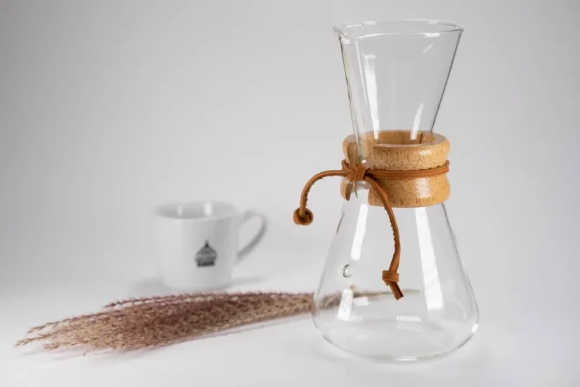 Glass Chemex with an elongated head, wooden handle, and leather string, white coffee cup, scattered coffee on a white table.