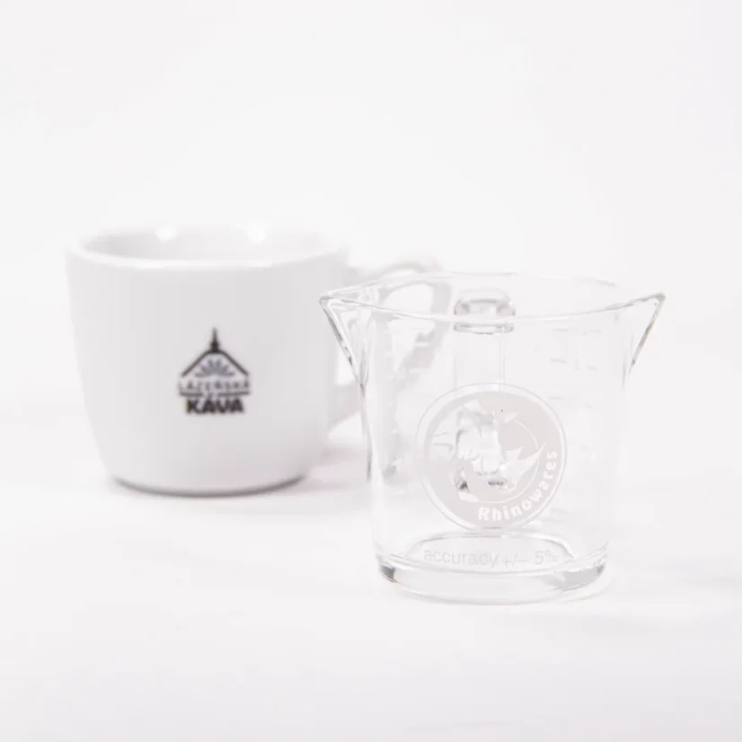 Sklenená odmerka na dvojité espresso pre baristov značky Rhinowares Double Spout Shot Glass vedľa šálky kávy.