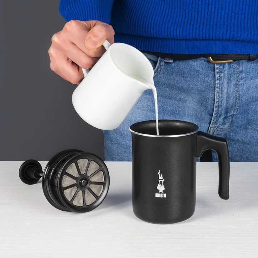 Milchaufschäumer in Schwarz mit einem Volumen von 166ml von Bialetti Tuttocrema und im Hintergrund ein Barista, der Milch in den Aufschäumer gießt.