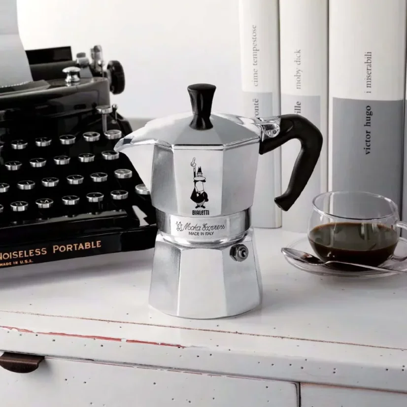 Silberne Bialetti Moka Express Kanne für 3 Tassen auf einer Küchenarbeitsplatte, im Hintergrund eine Schreibmaschine.