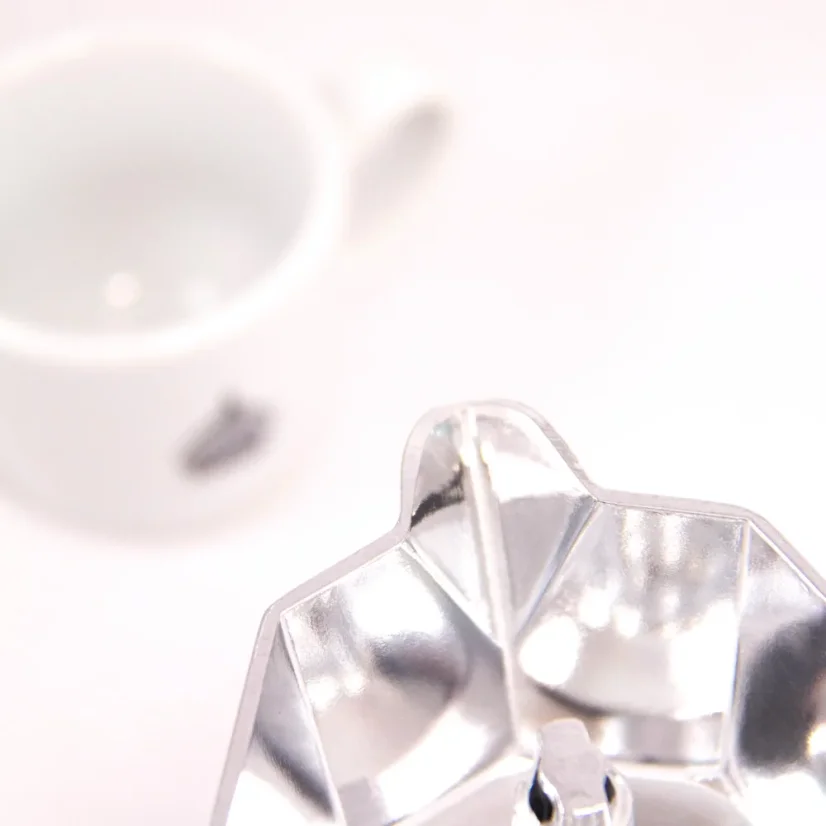 Detail of the spout of a silver Bialetti Moka Elettrika Standard coffee maker on a white table, accompanied by a cup with coffee-themed designs.