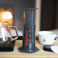 AeroPress Aerobie - plastmasas AeroPress uz koka galda ar filtru, balts krūzītis ar logotipu, stikla trauks ar rokturi, apgaismota telpa.