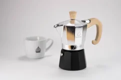 Silberne Moka Kanne Forever Miss Moka Woody mit Holzgriff auf weißem Tisch, zusammen mit einer Porzellantasse Kaffee. Silberne Moka Kanne Forever Miss Moka Woody mit Holzgriff auf weißem Tisch, zusammen mit einer Porzellantasse Kaffee.