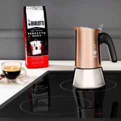 Bialetti New Venus Cooper kávéfőző indukciós lapon. A háttérben szemes kávé csomagolása, amely alkalmas erre a főzési módszerre.