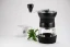 Black Hario Skerton Pro manual coffee grinder with a cup of coffee and a green plant.