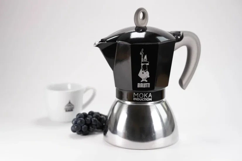 Cafetière moka en aluminium convenant à l'induction avec le logo du fabricant, la société italienne Bialetti, accompagnée d'une tasse comportant un fruit à baies.