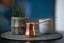Kupferne Kaffeekanne auf einer geflochtenen Tischmatte, im Hintergrund eine Zimmerpflanze im Blumentopf und eine Kerze.