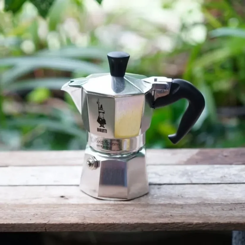 Bialetti Moka Express pot on a wooden bench in nature.