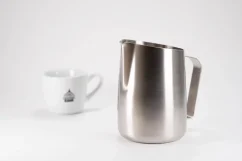 Stainless steel milk frothing jug on a white table together with a porcelain cup.