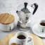 Silver moka pot for 3 cups of Bialetti Moka Express on a white table, accompanied by prepared coffee in porcelain cups and a white sugar bowl with a wooden lid.