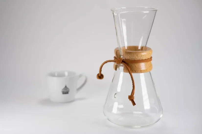 Glass Chemex with an elongated head, wooden handle, leather string, and a white coffee cup with a logo.