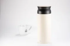 White stainless steel thermal bottle with a capacity of 500 ml on a white background with a cup of coffee