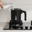 Pouring milk from a carafe into a Bialetti Tuttocrema 166ml milk frother placed on a gas stove.