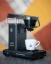 Moccamaster noir Cup One sur une table grise avec une tasse blanche sur un fond coloré.