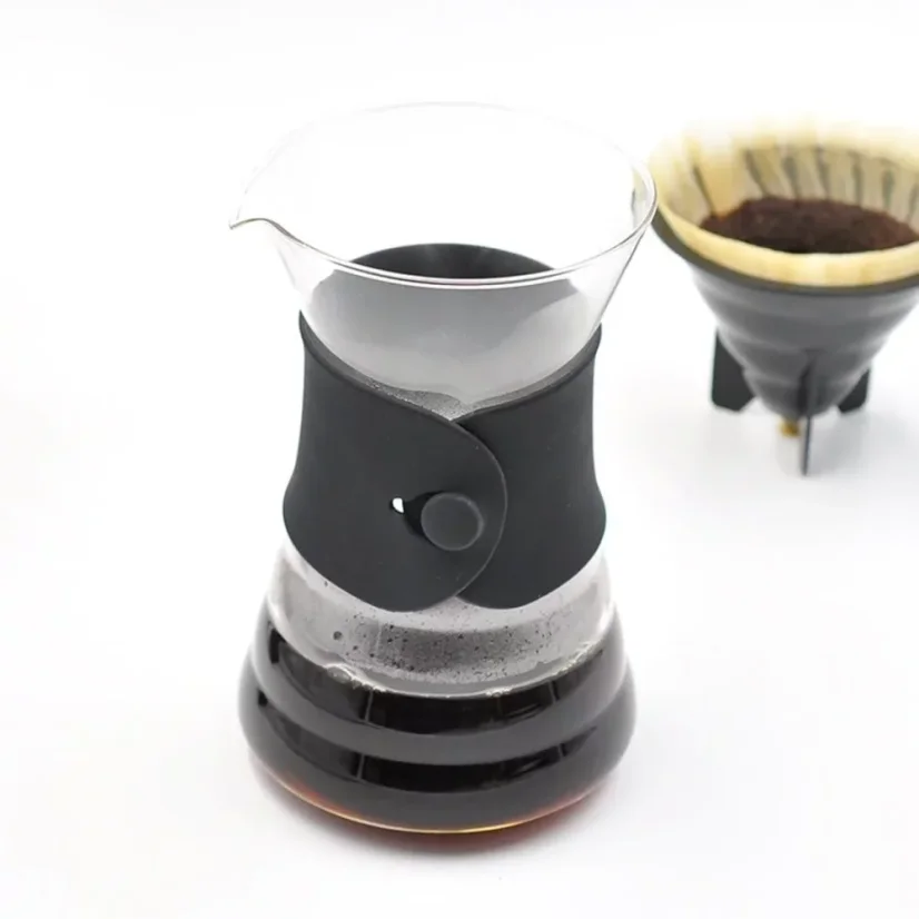 Hario V60 Drip Decanter s čiernou koženou rukoväťou, v pozadí papierový filter s mletou kávou.