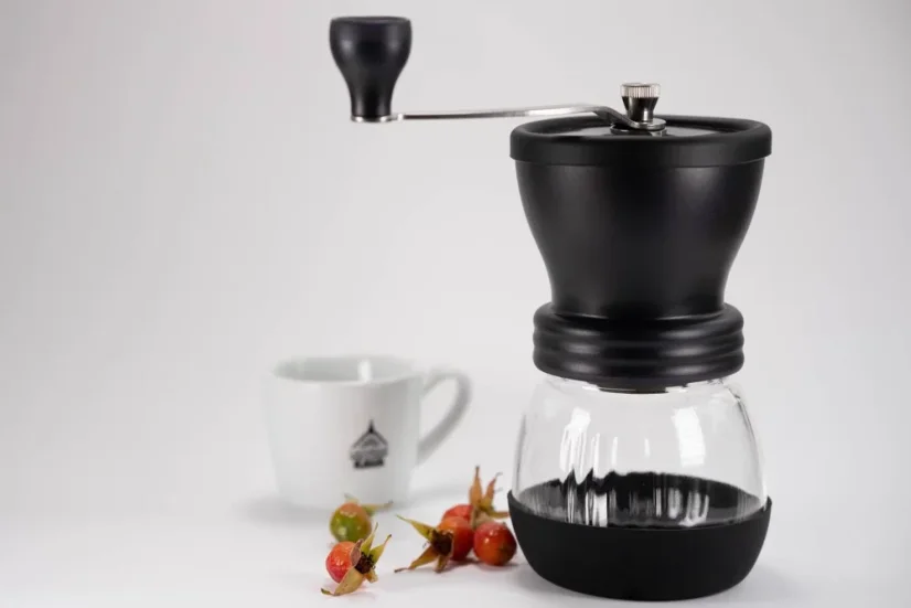 Hario Skerton plus čierny ručný mlynček na kávu so šípkami a v pozadí s hrnčekom kávy.