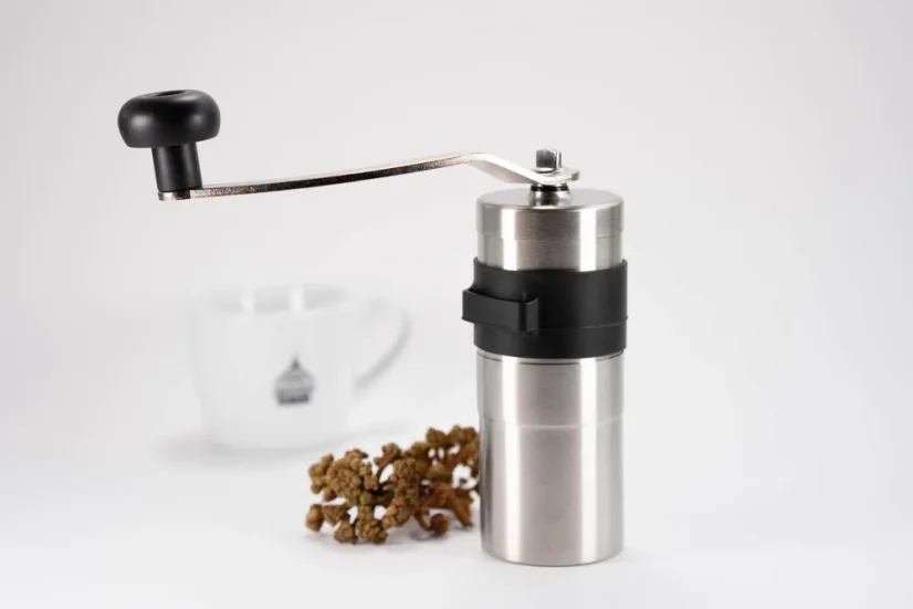 Silver manual coffee grinder on a white background with a cup of coffee and a sprig