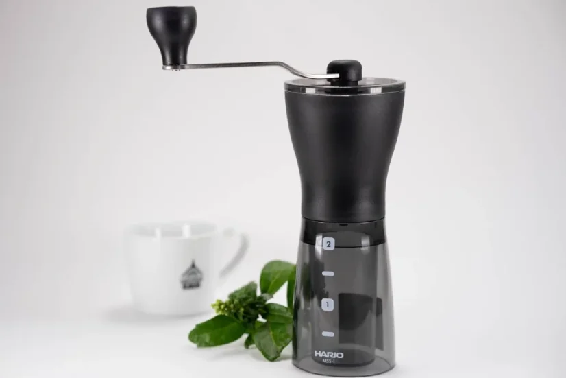 Čierny ručný mlynček na kávu na bielem stole s rastlinou a bielym šálkom na kávu v pozadí