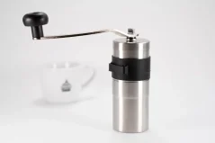 Silver manual coffee grinder on a white background with a cup of coffee