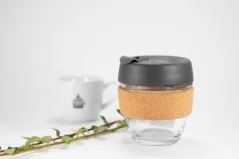 Glass thermal mug with a capacity of 227 ml from KeepCup Brew Cork Nitro with a grey lid and cork holder, situated on a white table with a branch, white mug with logo on a white background.