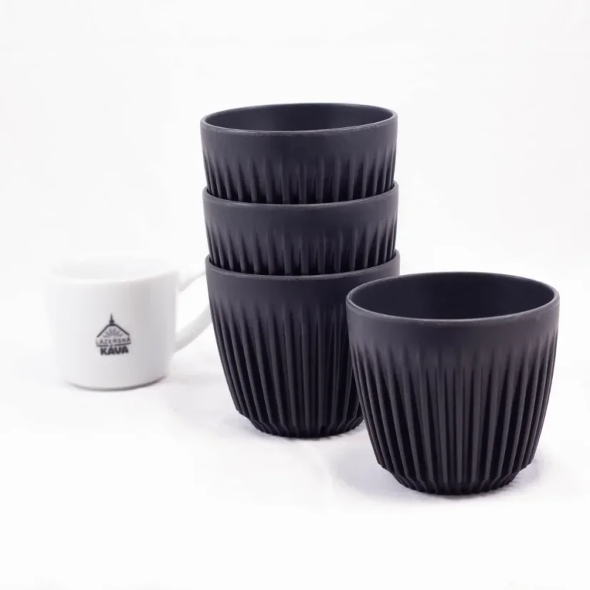 Ekologické termo hrnčeky v čiernej farbe značky Huskee Charcoal na bielej stolovej doske s porcelánovým šálkom na bielem pozadí