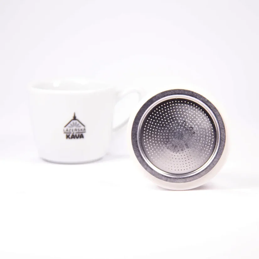Filtro argentato per caffettiera Moka Bialetti appoggiato su un bordo su sfondo bianco con una tazza di caffè.