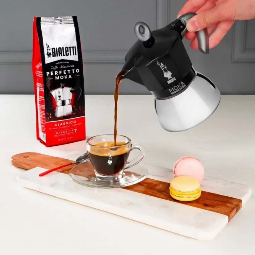 ukázka servírování kávy z hliníkové moka konvičky talianskej značky Bialetti do priesvitného šálku na drevenom podnosu s vrecúškom kávy a malými makronkami