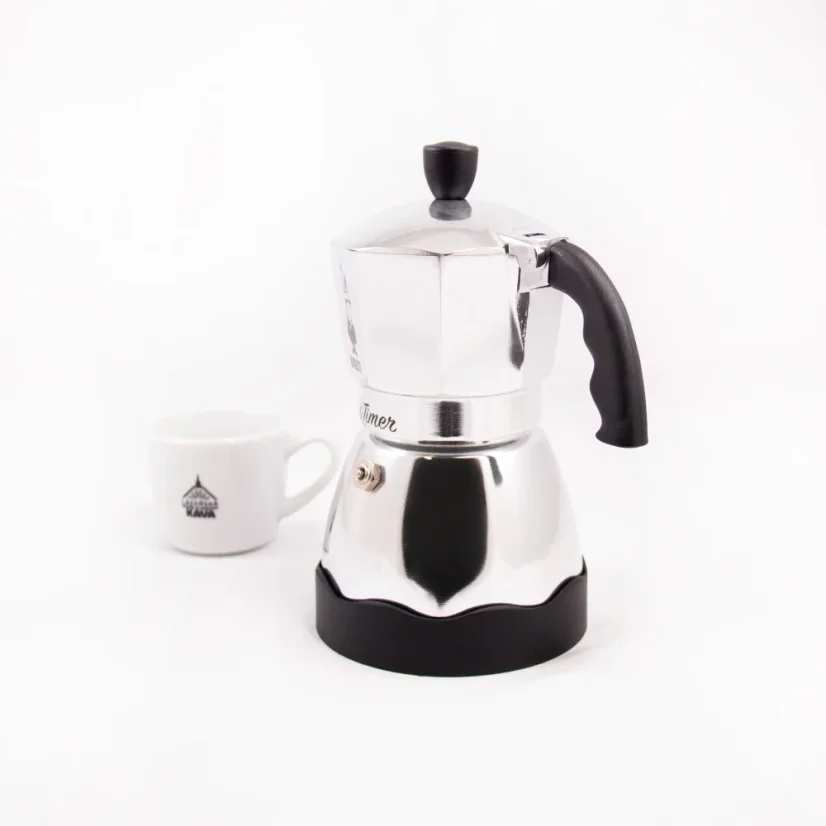 Silver Bialetti Moka Timer coffee maker for 3 cups, rear view, with a cup of coffee in the background.
