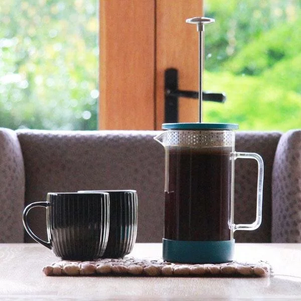 Proces prípravy kávy v modrom French press na bielem stole s dvoma čiernymi šálkami na kávu s výhľadom do prírody.