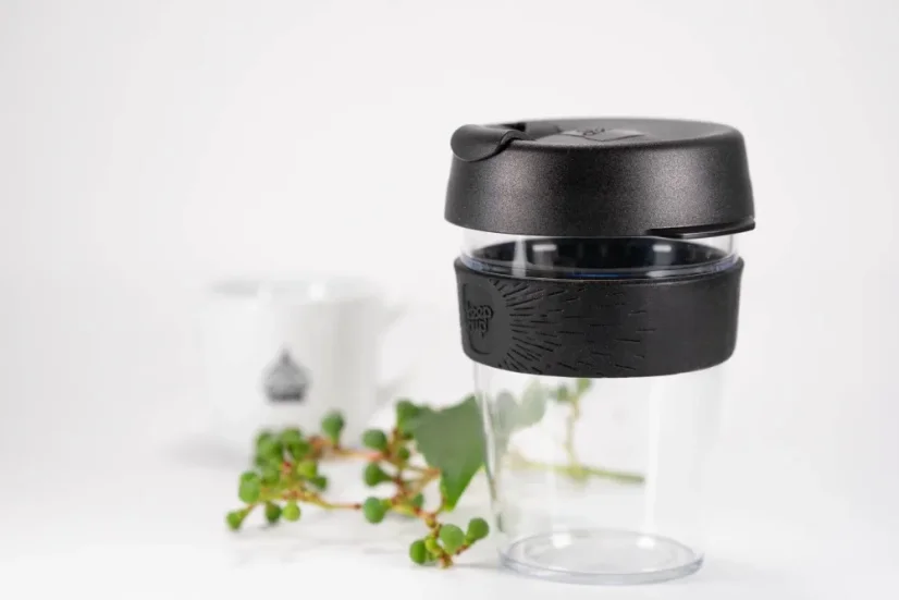 Plastic thermal mug with a rubber holder on a white table with plants and a white mug with a logo.