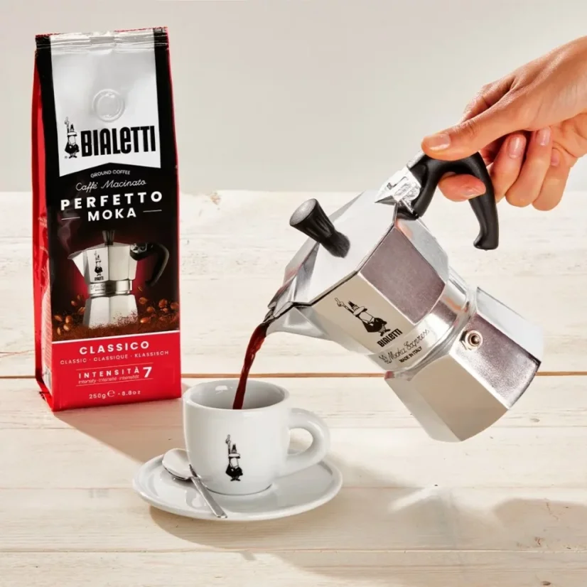 Pouring coffee prepared in a Bialetti Moka Express into a white cup with a logo.