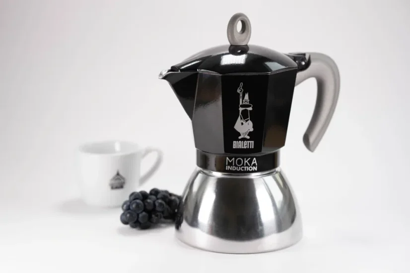 Aluminum moka pot suitable for induction with the logo of the Italian manufacturer Bialetti, displayed alongside a cup with coffee beans and berries.