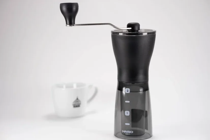 Čierny ručný mlynček na kávu na bielem stole a biely hrnček na kávu v pozadí.