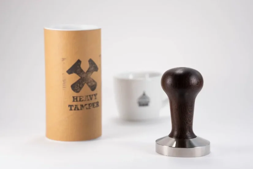 Heavy Temper with a diameter of 58 mm and a black handle on a white table with a cup of coffee and the original packaging for the temper.